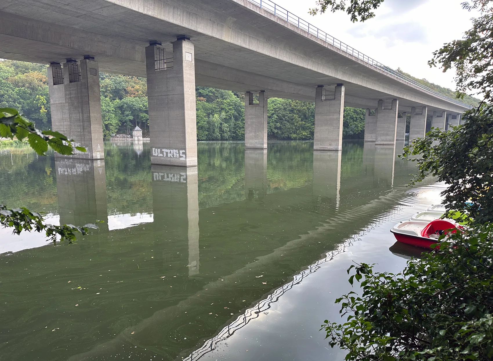 Betonbrücke über dem Seilersee mit Spiegelungen der Pfeiler im Wasser und einem kleinen rot-weißen Boot am Ufer.
