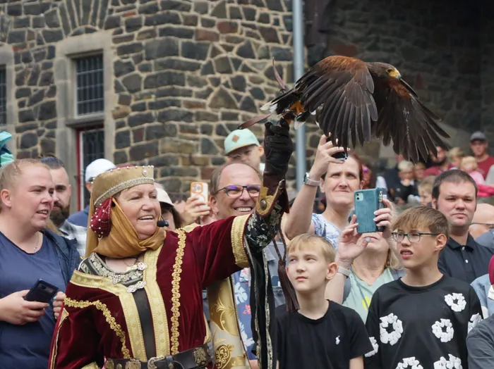 Eine Frau in historischem Gewand präsentiert einen Greifvogel mit ausgebreiteten Flügeln vor einer Zuschauermenge.