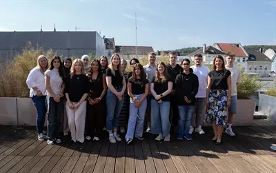 Eine Gruppe von 18 Auszubildenden und zwei Ausbildungsleiterinnen posiert für ein Gruppenfoto auf einer Holzterrasse vor einer Stadtkulisse.