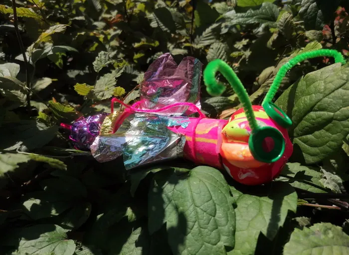 Ein buntes, handgefertigtes Kunstwerk in Form eines Insekts aus glänzender Folie und bunten Materialien liegt auf grünen Blättern.
