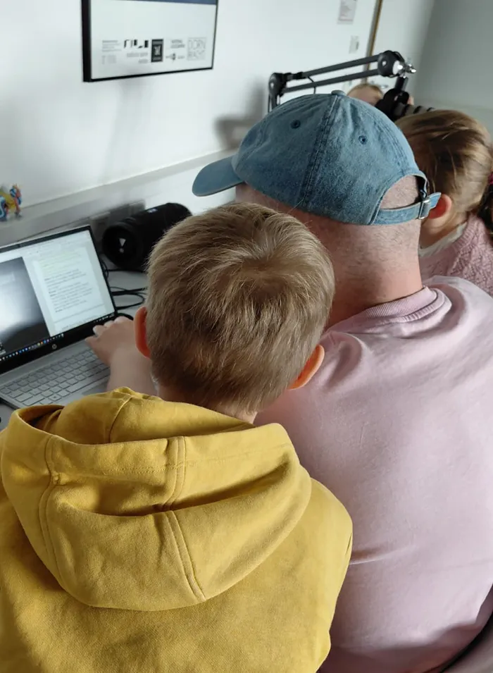 Ein Mann mit blauer Kappe und ein Kind in einem gelben Hoodie blicken gemeinsam auf einen Laptop-Bildschirm.