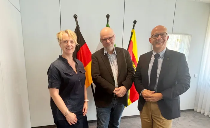 Leslie Kamphuis, Dr. Michael Achenbach und Landrat Ralf Schwarzkopf stehen nebeneinander vor Flaggen.