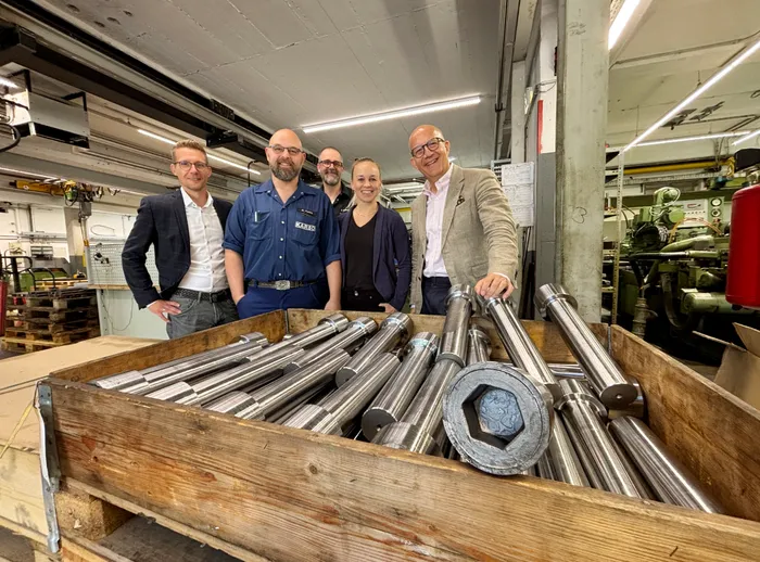 Fünf Personen stehen in einer Werkstatt hinter einer Holzkiste, die mit großen, metallischen, zylindrischen Industrieteilen gefüllt ist.