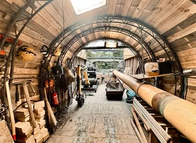 Innenansicht eines hölzernen Stollens unter Konstruktion mit Metallbögen, Baumaschinen und Werkzeugen, der auf einen hellen Ausgang zuläuft.