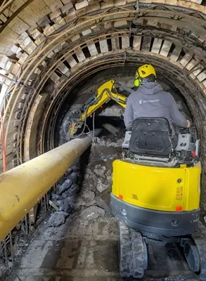 Ein Arbeiter bedient einen kleinen gelben Bagger in einem mit Holz und Metall verstärkten Tunnelstollen.