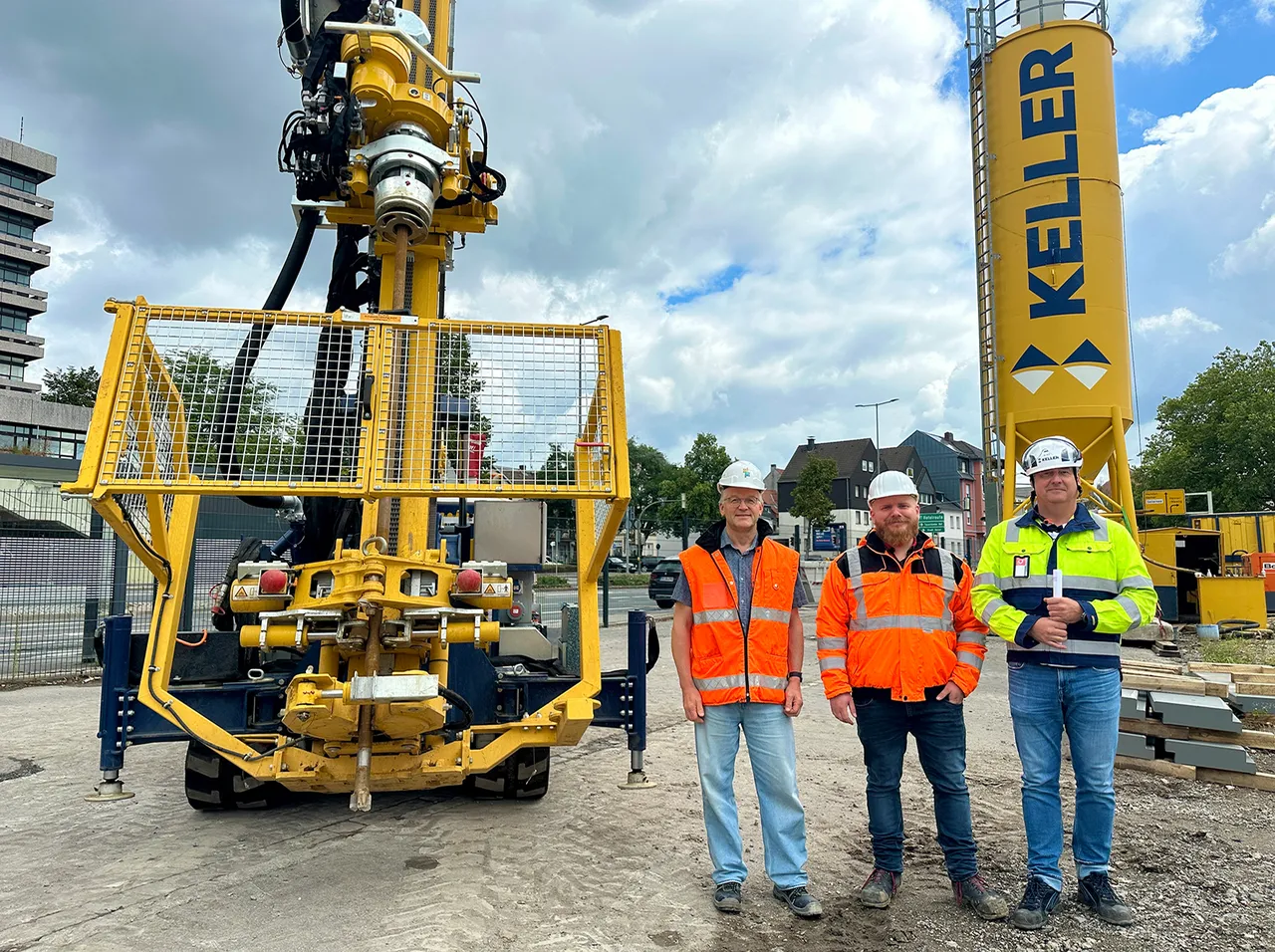 Drei Männer in Warnwesten und Schutzhelmen stehen vor einem großen gelben Bohrgerät und einem gelben Keller-Silo auf einer Baustelle.