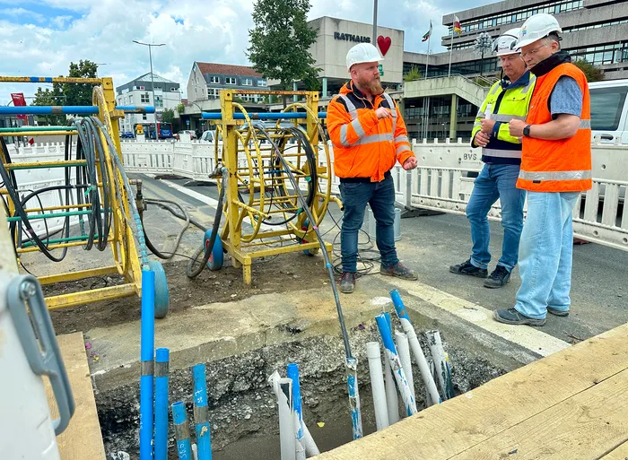 Drei Männer in Warnwesten und Schutzhelmen stehen an einer Baustelle mit einem offenen Schacht und gelbem Bohrgerät vor dem Rathaus.