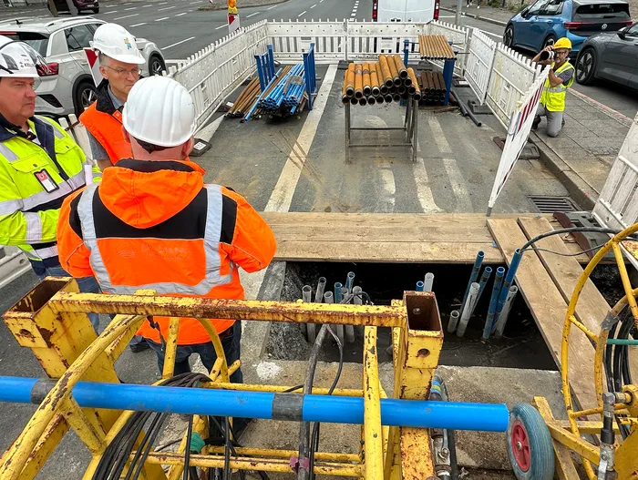 Bauarbeiter in Warnkleidung und Schutzhelmen blicken in einen offenen Schacht auf einer Baustelle.