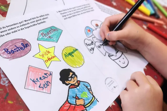 Kinderhände malen mit einem schwarzen Stift in ein buntes Arbeitsblatt mit Begriffen wie „Basteln“ und „Phantasie“ sowie einer Superhelden-Illustration.