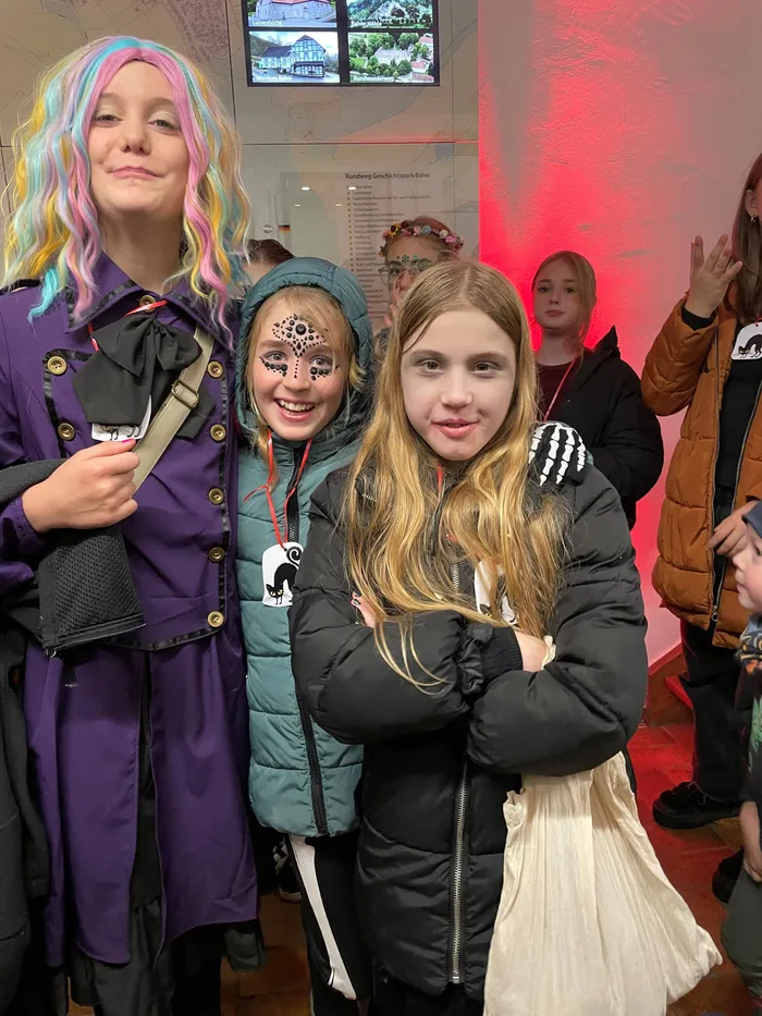 Vier Kinder in Halloween-Kostümen und mit Gesichtsbemalung posieren gemeinsam vor einer rot beleuchteten Wand.