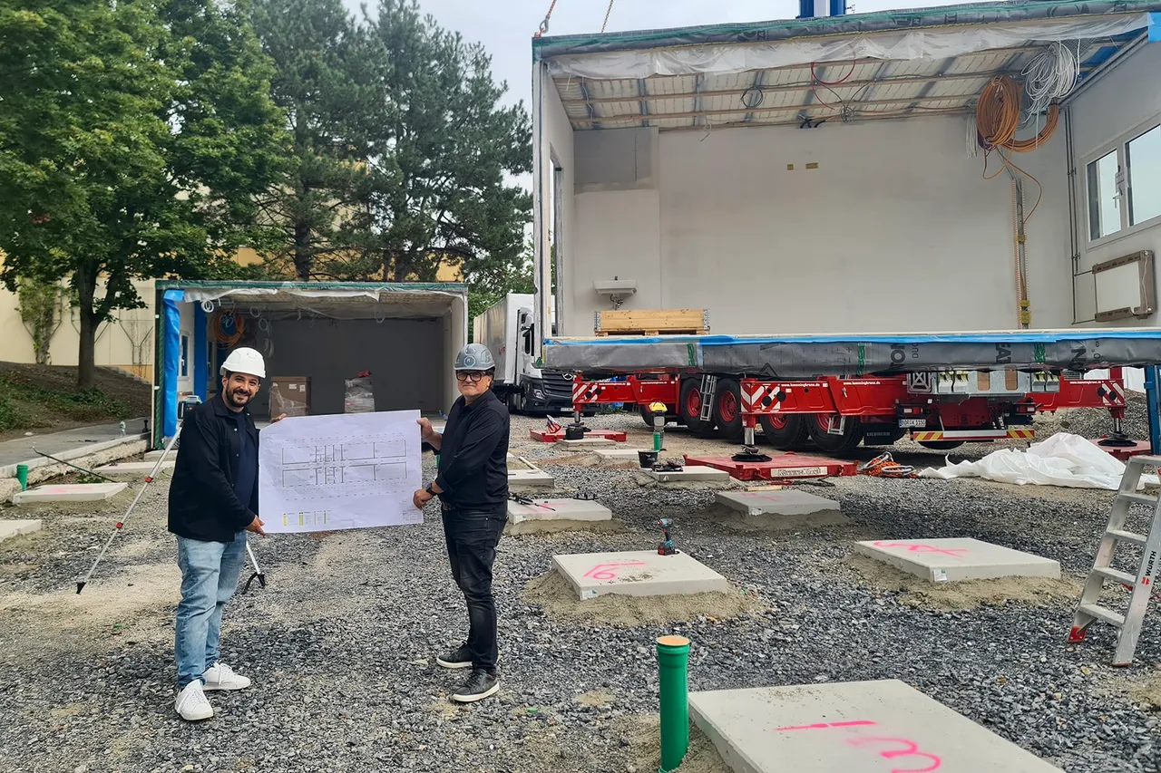 Zwei Männer halten einen Bauplan vor einem weißen Container auf einem roten LKW-Anhänger auf einer Baustelle.