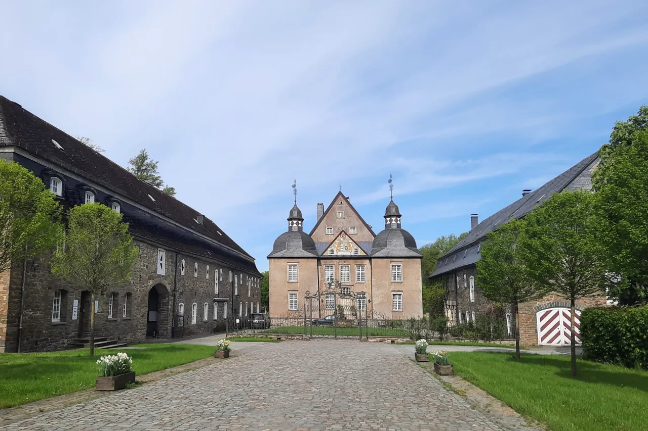Das Wasserschloss Neuenhof mit zwei Türmen, flankiert von zwei langgestreckten Steingebäuden und einem gepflasterten Weg im Vordergrund.