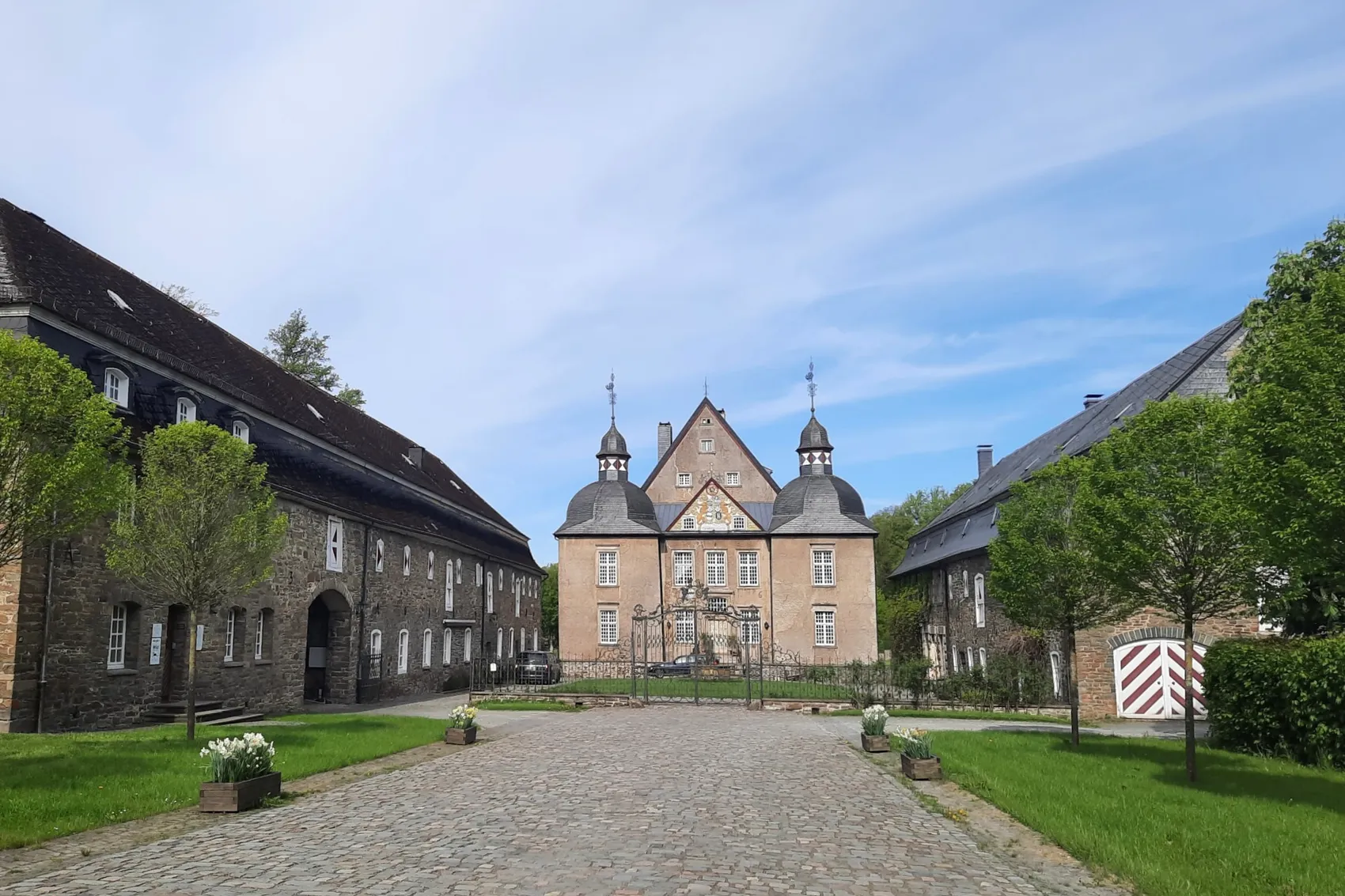 Das Wasserschloss Neuenhof mit zwei Türmen, flankiert von zwei langgestreckten Steingebäuden und einem gepflasterten Weg im Vordergrund.