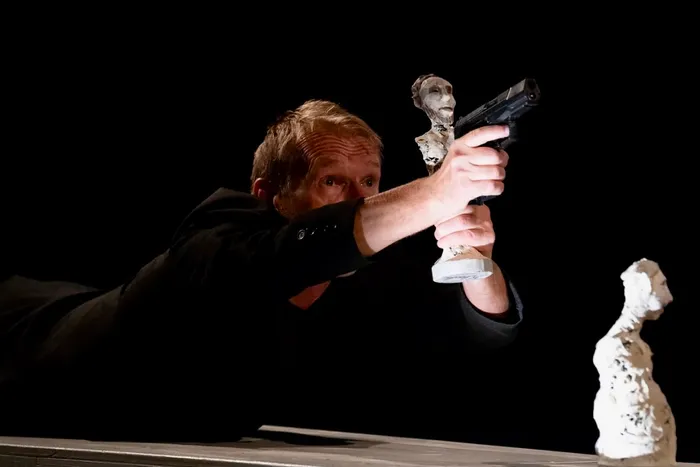 Ein Mann in schwarzer Kleidung zielt während einer Theateraufführung mit einer Pistole auf eine weiße Skulptur.