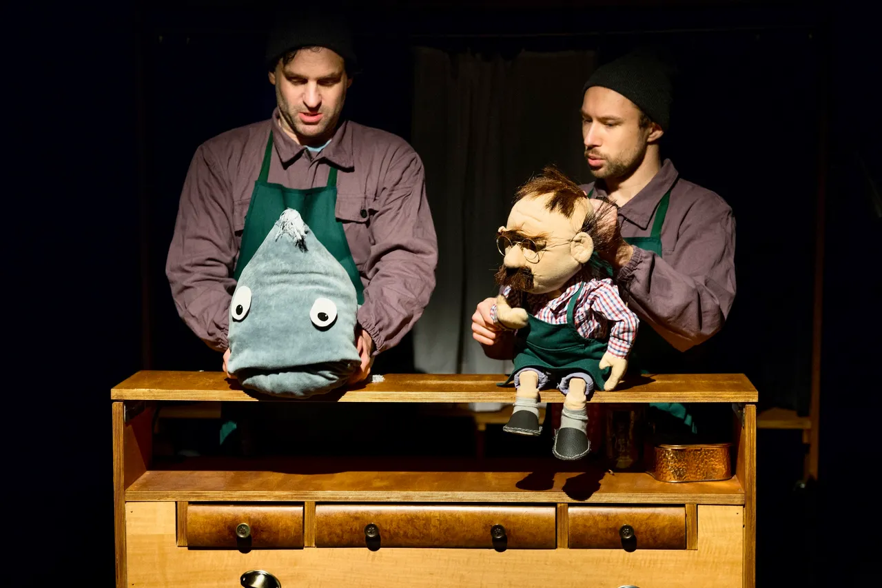 Zwei Puppenspieler halten eine große graue Fischpuppe und eine menschliche Puppenfigur vor einem Holzschrank.