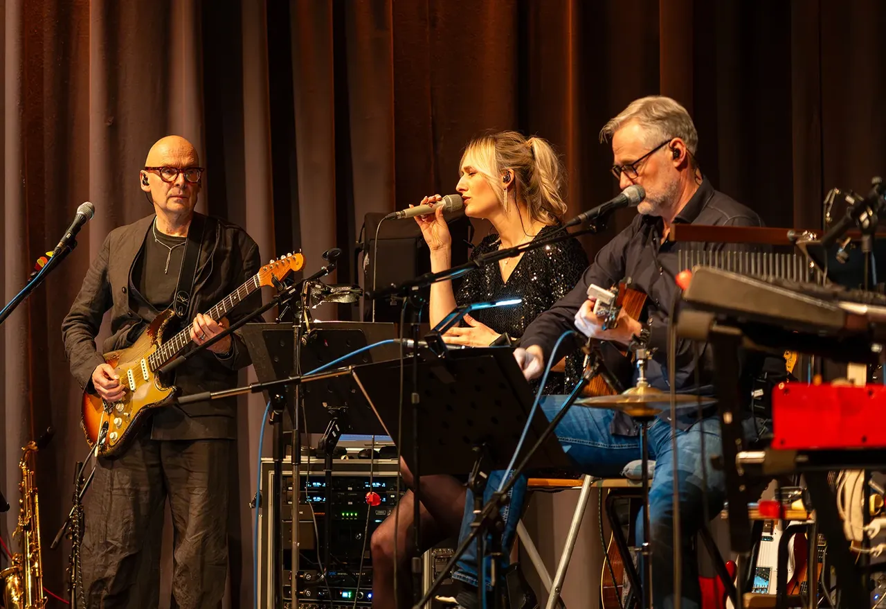 Die Band „Good Wood“ bei einem Live-Auftritt mit einem E-Gitarristen, einer Sängerin und einem Akustikgitarristen vor einem dunklen Vorhang.
