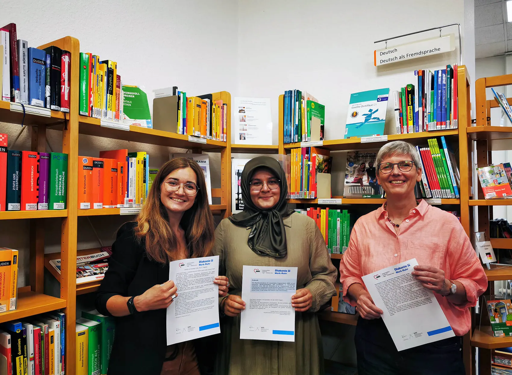 Drei Frauen halten Dokumente vor Bücherregalen mit Büchern zum Thema Deutsch als Fremdsprache hoch.