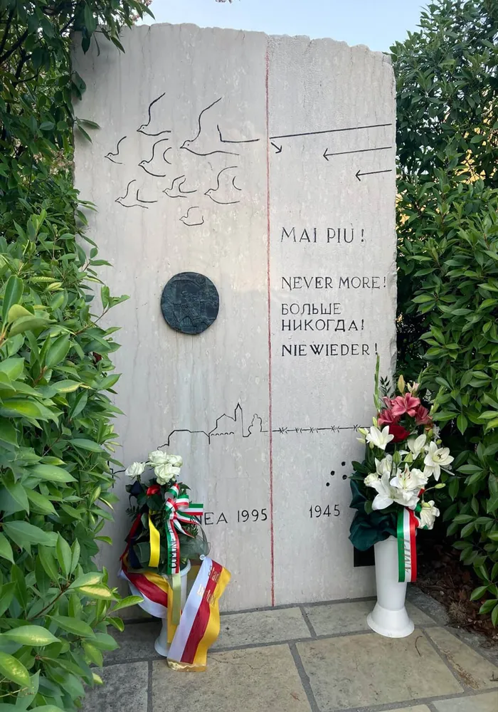 Ein Steindenkmal mit Vogel- und Stadtmotiven sowie dem mehrsprachigen Schriftzug „Never More!“. Am Fuß stehen Blumen mit deutschen und italienischen Bändern.