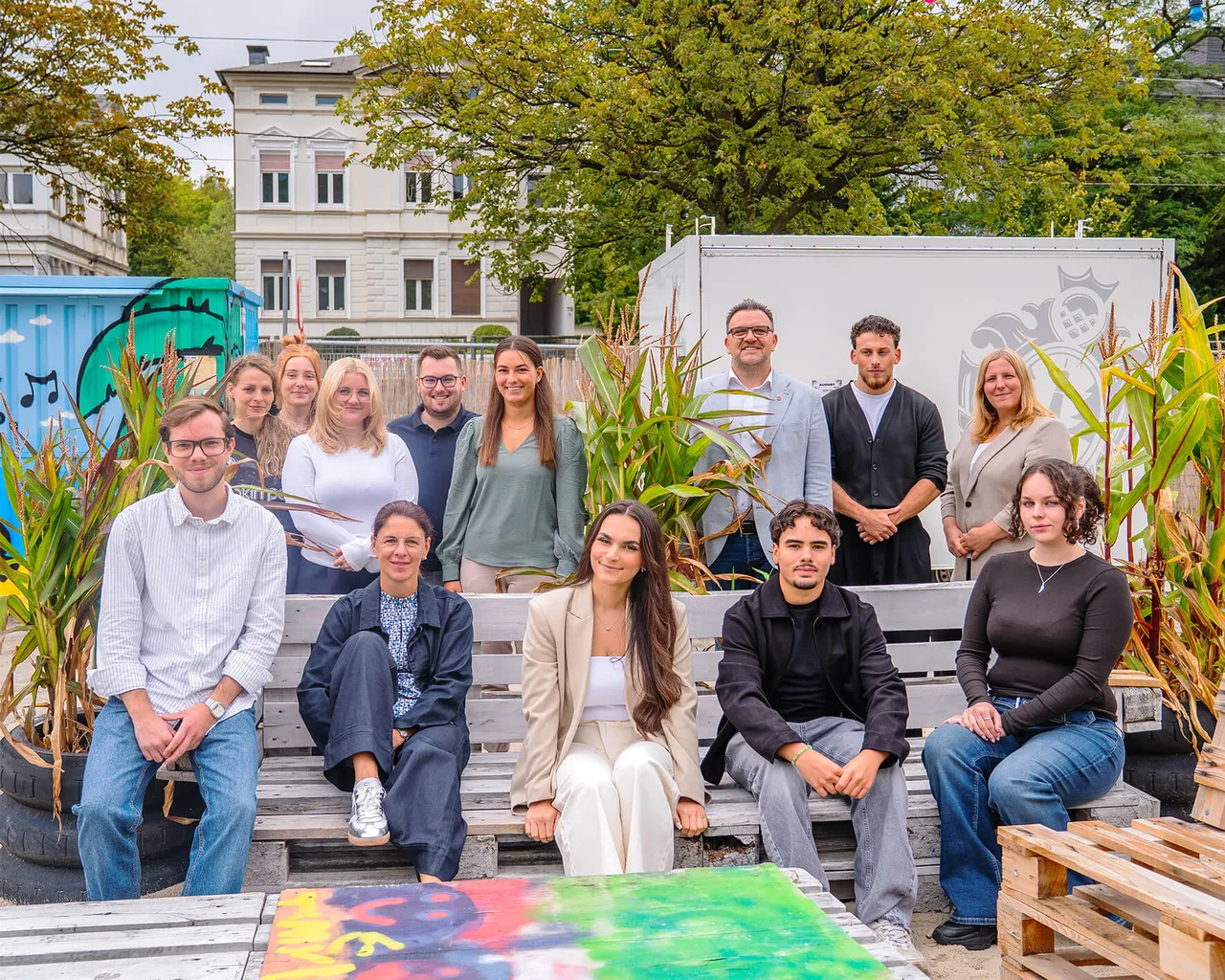 Dreizehn Personen, darunter Auszubildende und der Bürgermeister, posieren für ein Gruppenfoto im Freien vor Maispflanzen und einem weißen Gebäude.