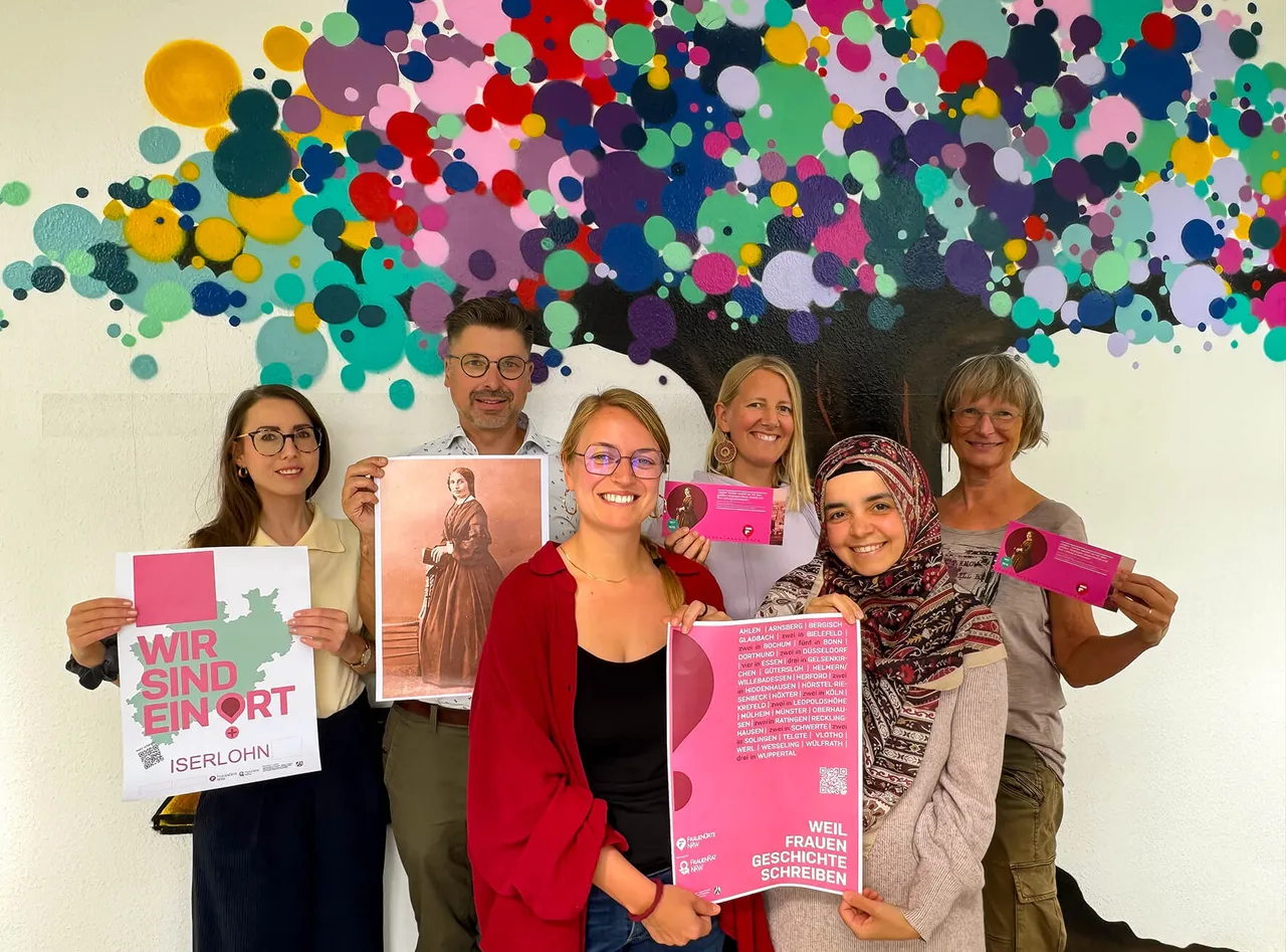 Sechs Personen posieren vor einer bunten, gepunkteten Wand und halten Plakate mit den Aufschriften „Wir sind ein Ort Iserlohn“ und „Weil Frauen Geschichte schreiben“.