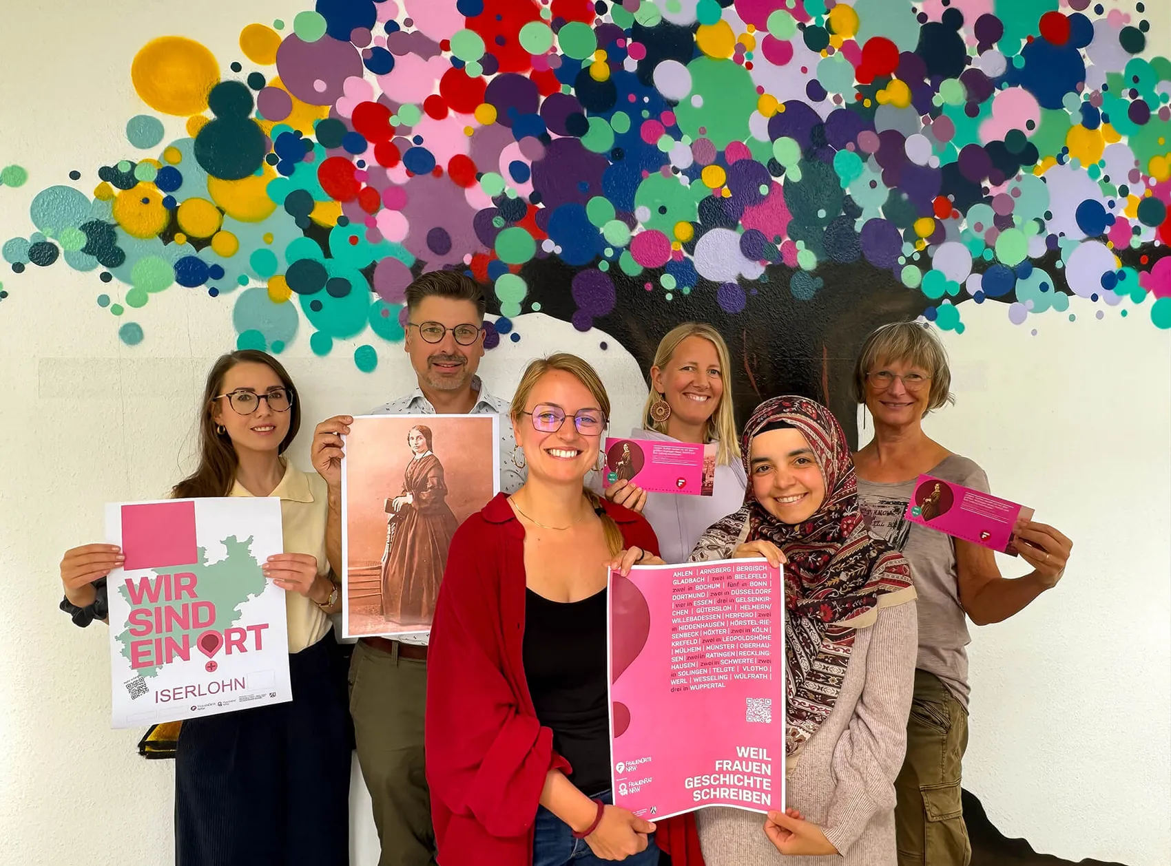 Sechs Personen präsentieren vor einem bunten, abstrakten Wandbild Plakate und Flyer zum Projekt „FrauenOrt“ in Iserlohn.