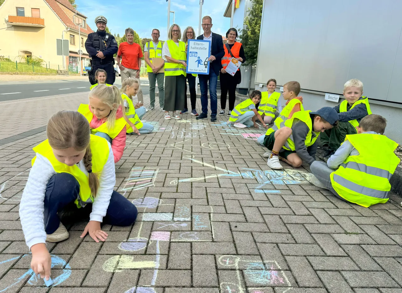 Kinder in gelben Warnwesten zeichnen mit Kreide auf den Boden, während im Hintergrund Erwachsene ein Schild für die neue „Eltern-Haltestelle THS“ halten.