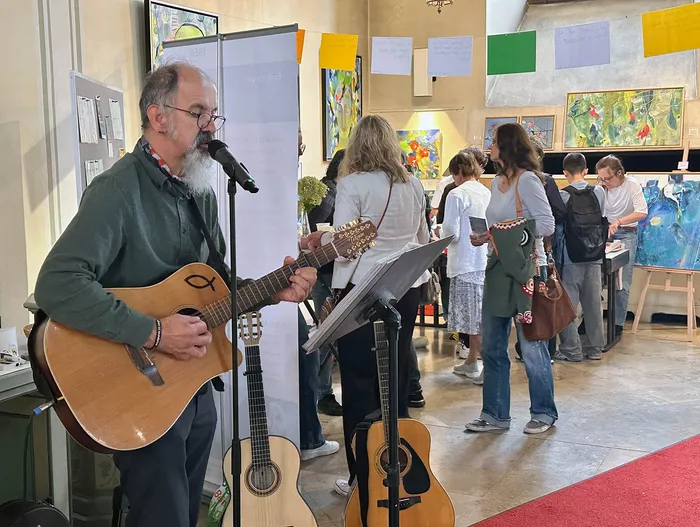 Liedermacher Lukas spielt Gitarre und singt in ein Mikrofon vor einer Kulisse aus Gemälden und Besuchern.