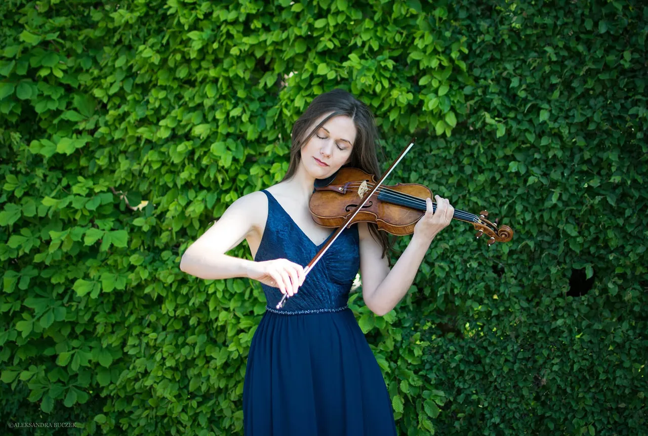 Lena Neudauer spielt in einem dunkelblauen Kleid mit geschlossenen Augen Geige vor einer grünen Hecke.