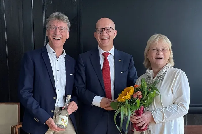 Rolf Klostermann (links) mit einer Flasche, Ralf Schwarzkopf (Mitte) und Dr. Christiane Todrowsky (rechts) mit einem Blumenstrauß lächeln gemeinsam.