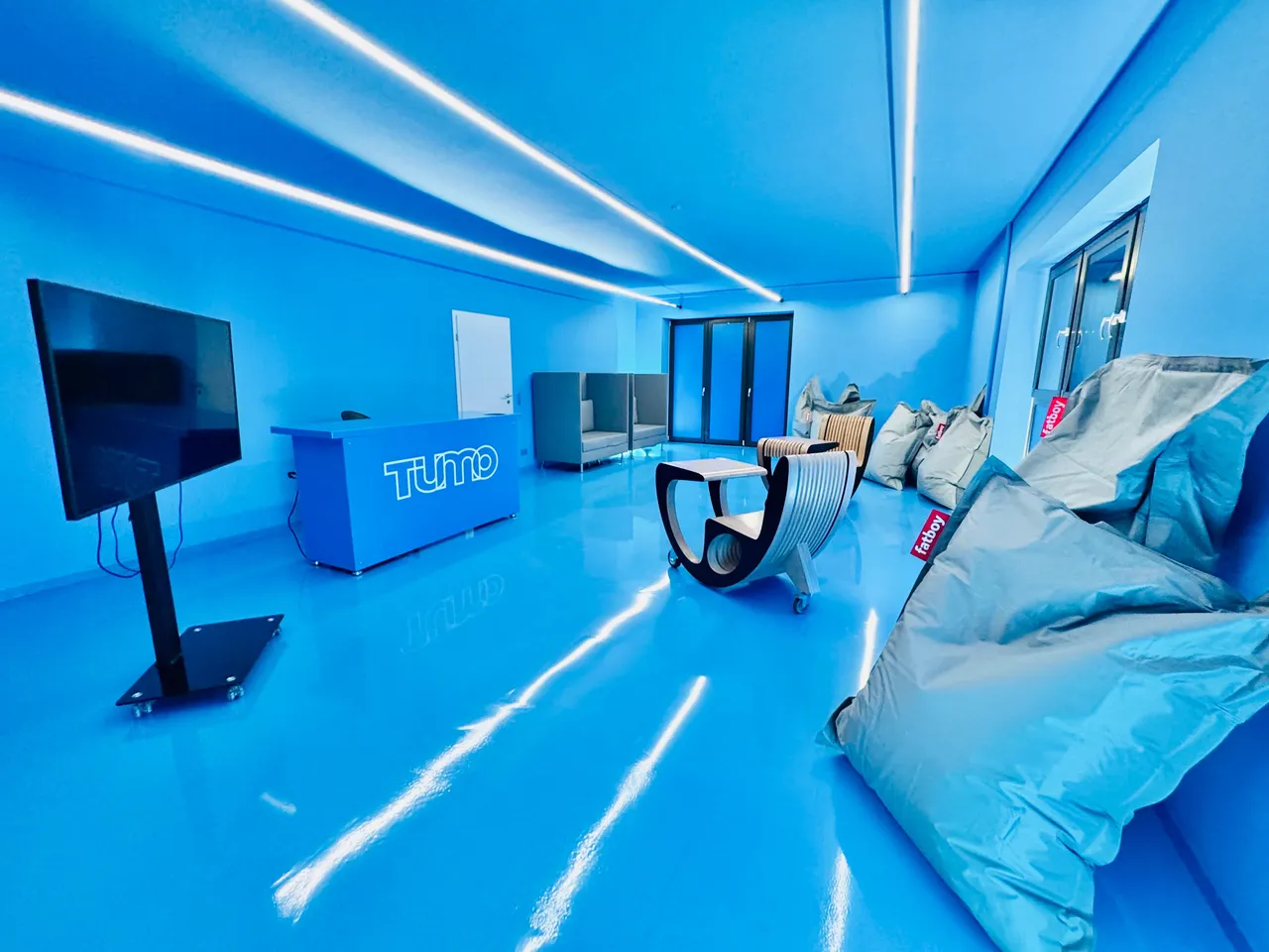 Heller, moderner Raum mit blauen Wänden und Boden, ausgestattet mit Sesselsäcken, einem Schaukelstuhl und einem Fernseher an der Wand. Im Hintergrund ist das Logo „Tumo“ zu sehen.