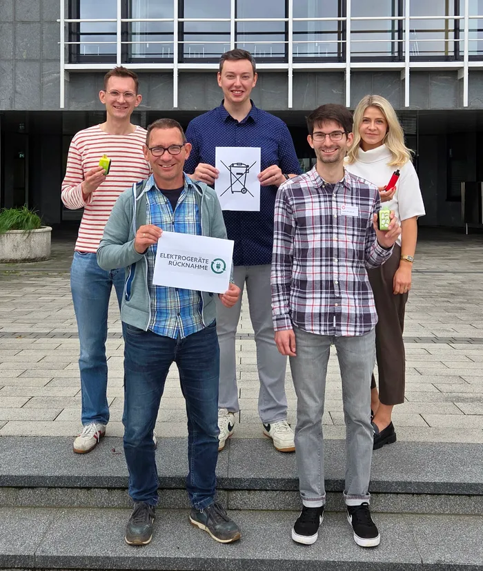 Fünf Personen halten Schilder mit einem durchgestrichenen Mülltonnen-Symbol und der Aufschrift „ELEKTROGERÄTE RÜCKNAHME“ sowie E-Zigaretten hoch.