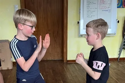 Zwei Jungen stehen sich in einem Raum gegenüber und halten ihre Hände für eine Übung erhoben.