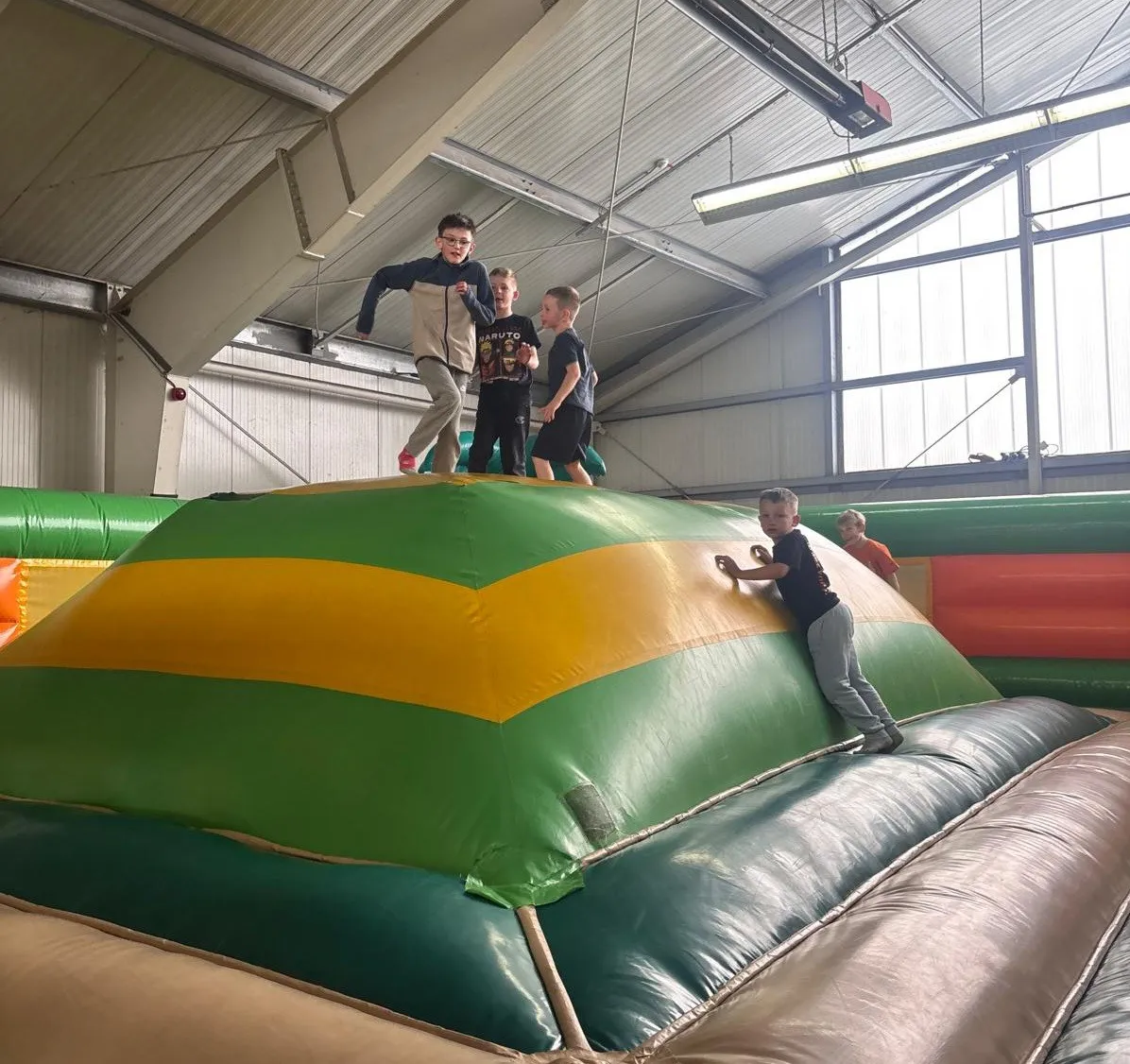 Mehrere Kinder und Jugendliche spielen auf einer großen, farbigen Hüpfburg in einer Turnhalle. Einige klettern hoch, andere halten sich fest.