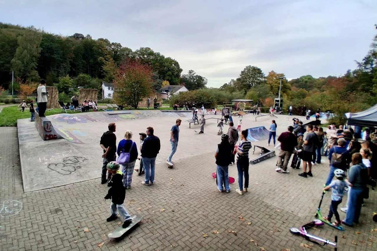 Menschen versammeln sich an einem Außen-Skatepark mit Betonrampen vor einer bewaldeten Kulisse.