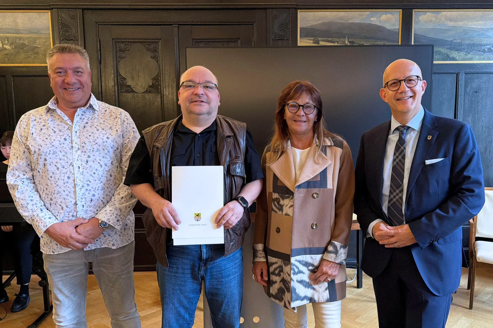 Vier lächelnde Personen stehen nebeneinander, wobei der Mann in der Mitte eine weiße Urkunde mit einem goldenen Wappen hält.