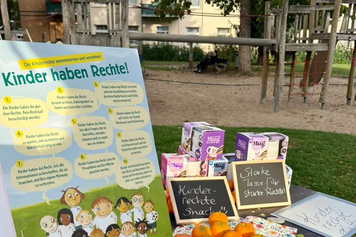 Ein Poster mit dem Titel „Kinder haben Rechte!“ steht auf einem Tisch neben Snacks und kleinen Kreidetafeln vor einem Spielplatz.