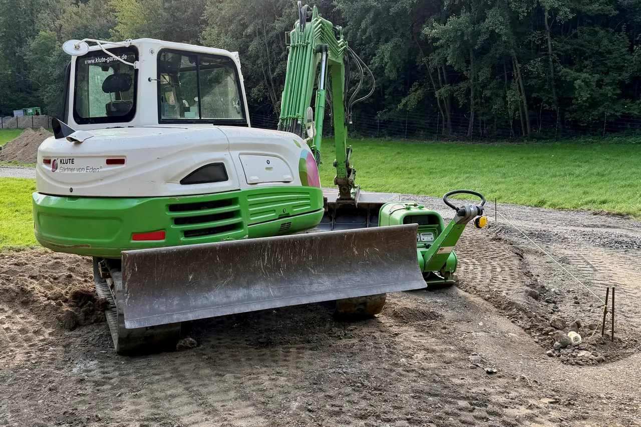Ein weiß-grüner Kettenbagger mit Frontschild bei Erdarbeiten auf einem unbefestigten Weg am Waldrand.