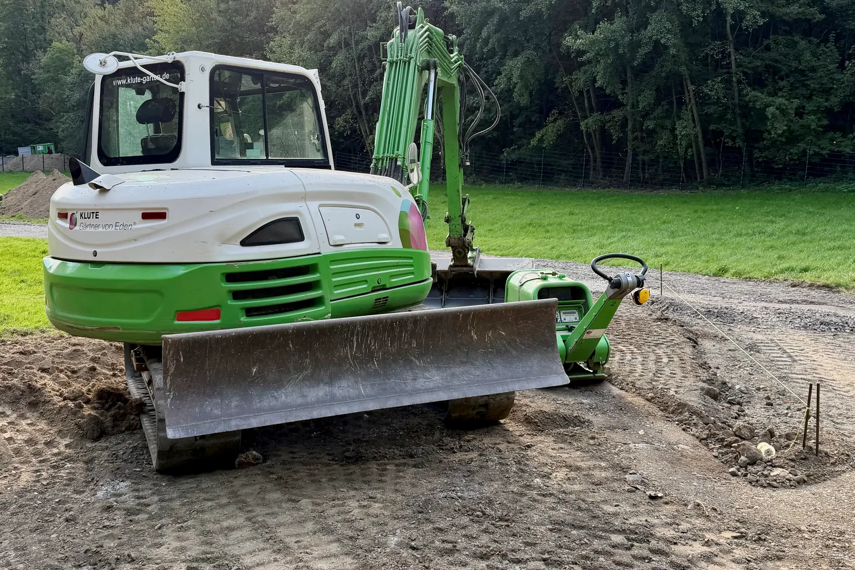 Ein weiß-grüner Kettenbagger mit Frontschild bei Erdarbeiten auf einem unbefestigten Weg am Waldrand.