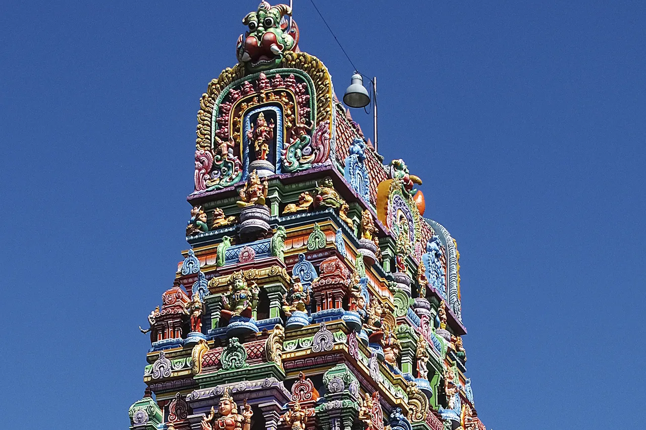 Ein farbenfroher, reich verzierter hinduistischer Tempelturm mit zahlreichen kleinen Götterfiguren vor blauem Himmel.