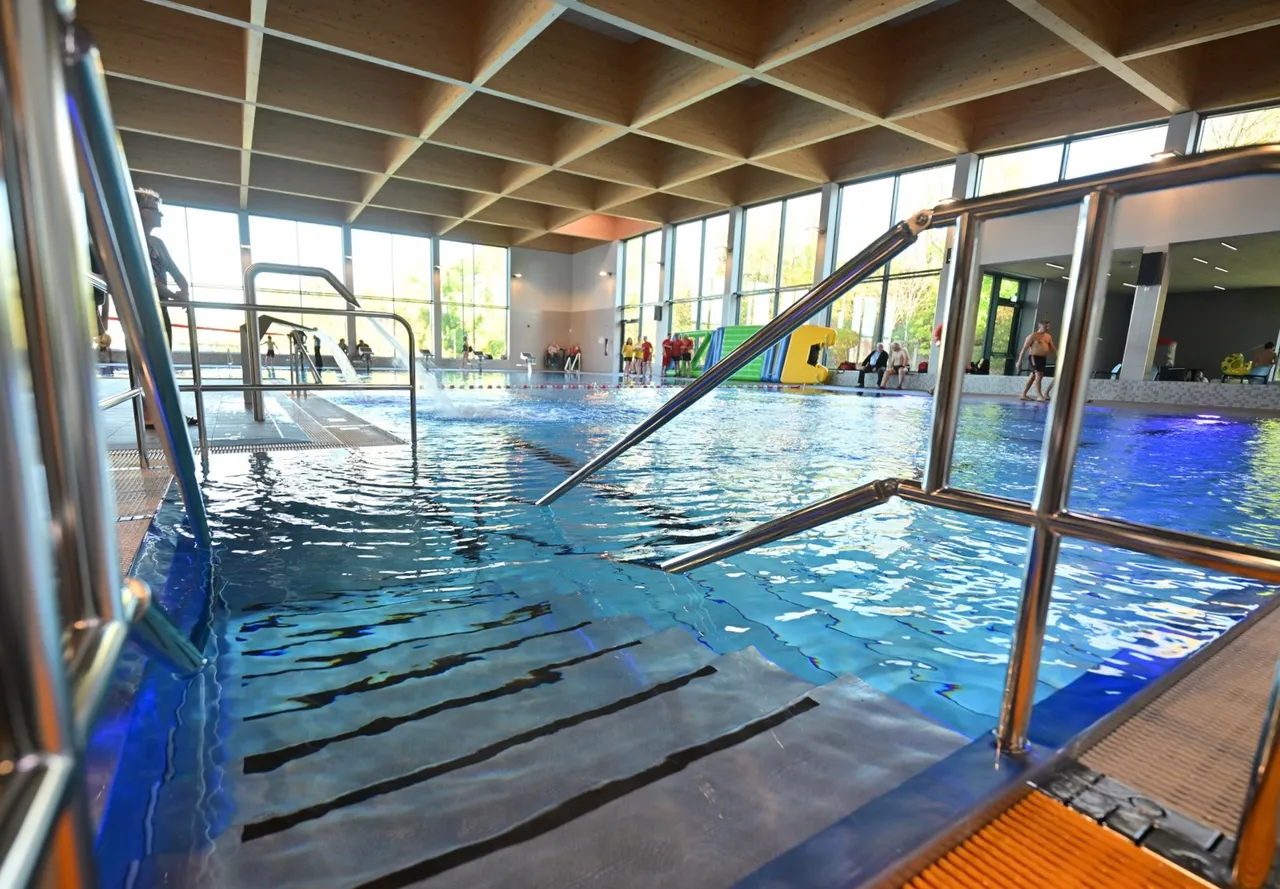Innenansicht des Hallenbads Felsenmeerbad mit blauem Wasser, Metallgeländern und großen Fensterfronten unter einer Holzdecke.