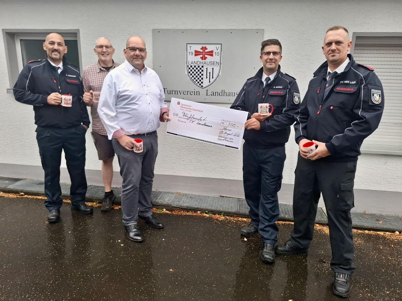 Fünf Männer, darunter Feuerwehrleute in Uniform, halten gemeinsam einen symbolischen Scheck über 500 Euro vor dem Gebäude des Turnvereins Landhausen.