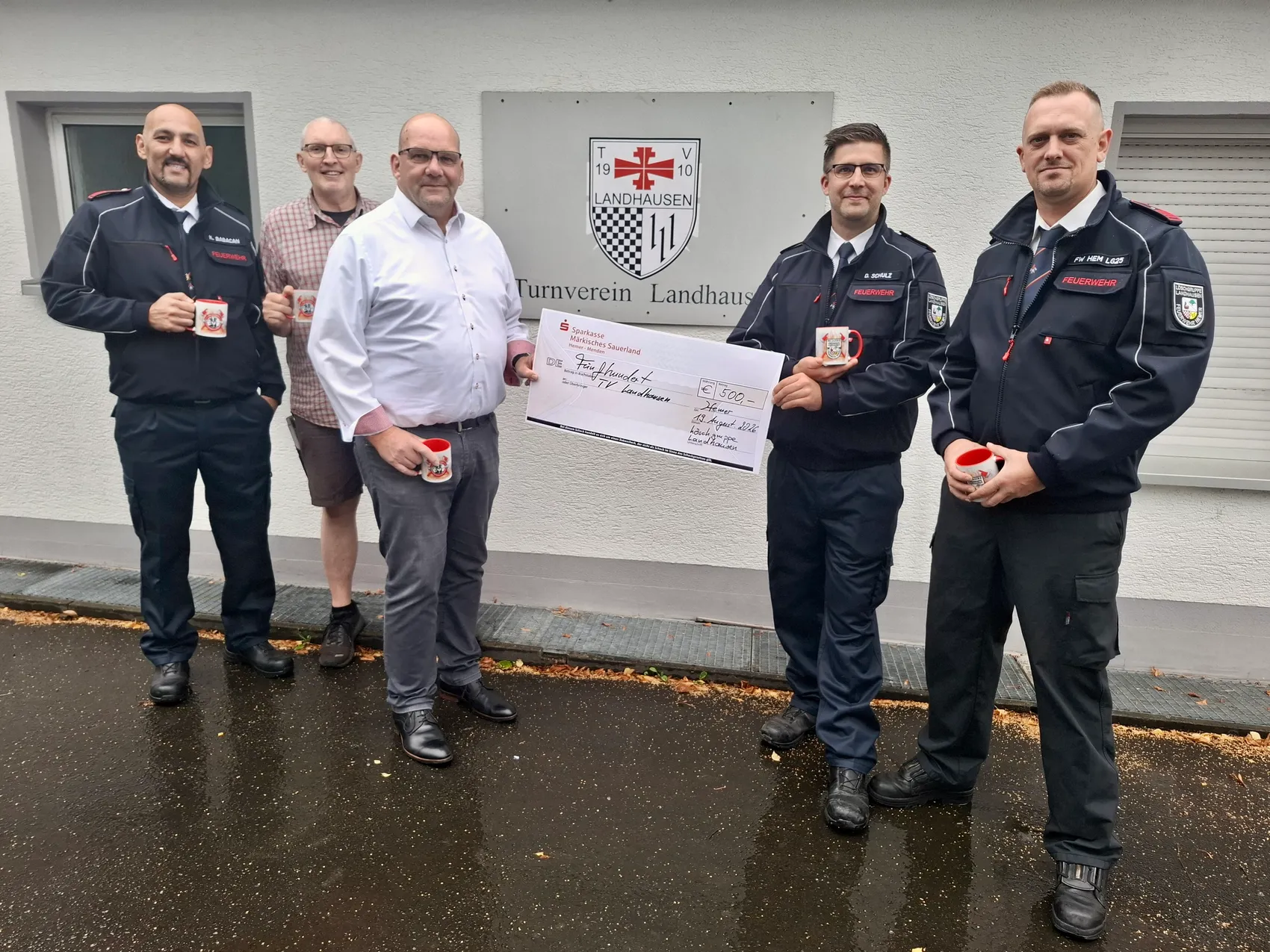 Fünf Männer, darunter Feuerwehrleute in Uniform, halten gemeinsam einen symbolischen Scheck über 500 Euro vor dem Gebäude des Turnvereins Landhausen.