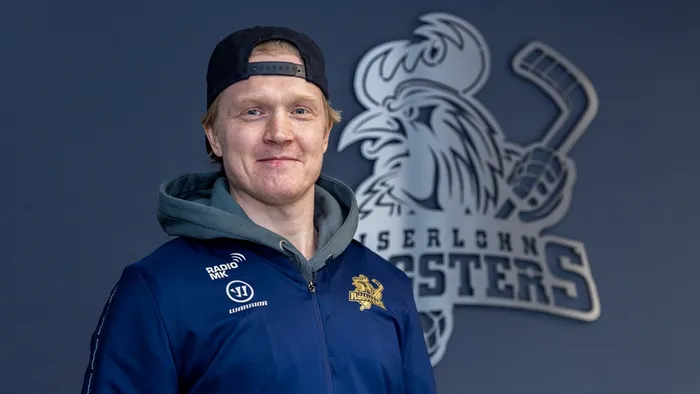 Porträt von Eishockeyspieler Emil Johansson mit Baseballkappe und Trainingsjacke der Iserlohn Roosters vor dem Vereinslogo.