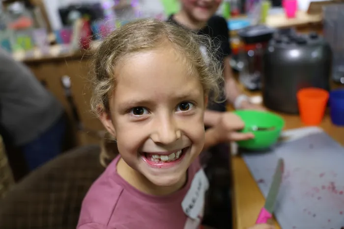 Ein lächelndes Mädchen mit blonden Locken blickt in die Kamera. Im Hintergrund sind andere Kinder und Küchenutensilien zu sehen.