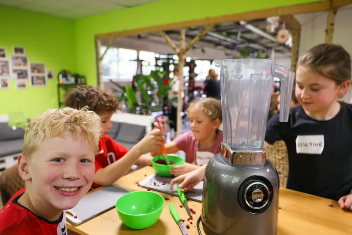 Kinder bereiten in einem hellen Raum gesunde Snacks zu. Ein Junge lächelt in die Kamera, während andere Kinder Beeren in Schüsseln füllen und einen Mixer bedienen.
