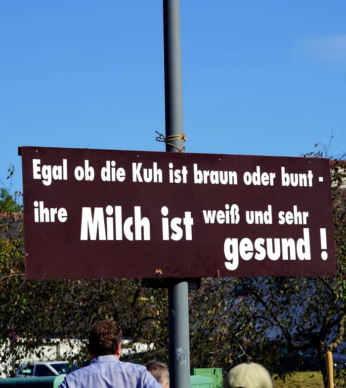 Braunes Schild mit der Aufschrift: „Egal ob die Kuh ist braun oder bunt - ihre Milch ist weiß und sehr gesund !“ vor blauem Himmel.