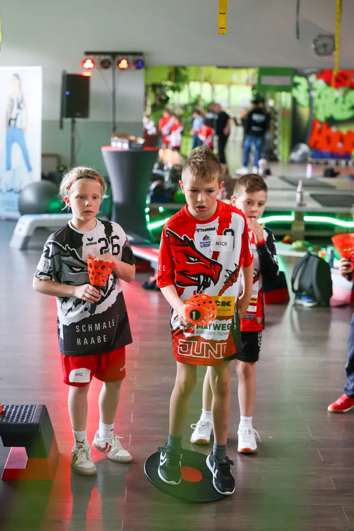Drei junge Handballspieler in Sportkleidung stehen nebeneinander in einer Halle. Der mittlere Spieler trägt ein rotes Trikot mit Drachenmotiv und die Aufschrift 