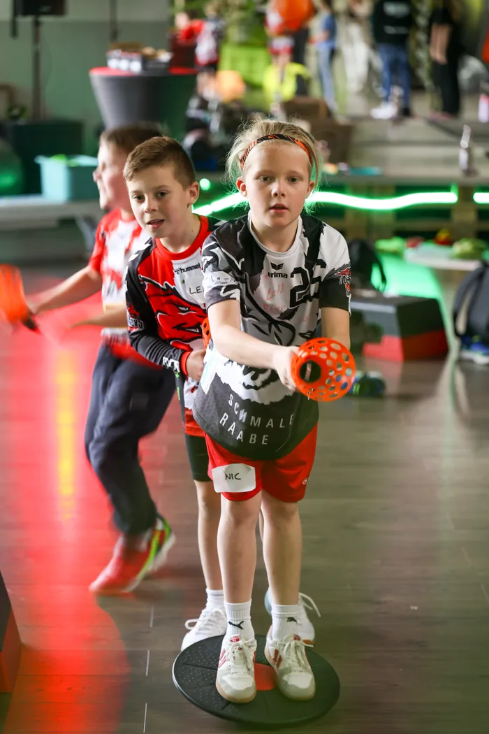 Ein Kind in Sportkleidung steht auf einer Drehscheibe und hält einen orangefarbenen Ball mit Griff. Im Hintergrund sind weitere Kinder zu sehen, die ebenfalls Sportkleidung tragen.