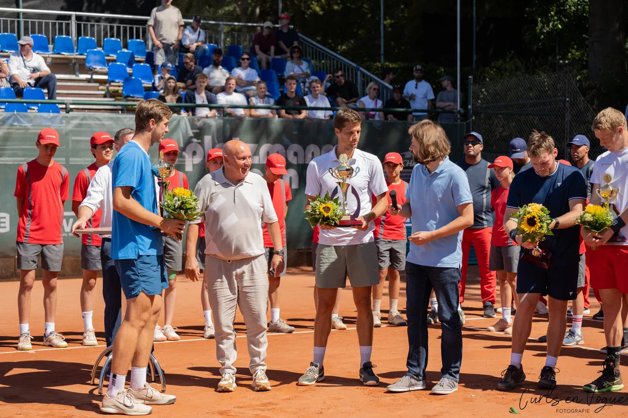 Ein Tennisspieler in einem weißen T-Shirt hält eine Trophäe und einen Sonnenblumenstrauß während einer Preisverleihung auf einem Sandplatz.