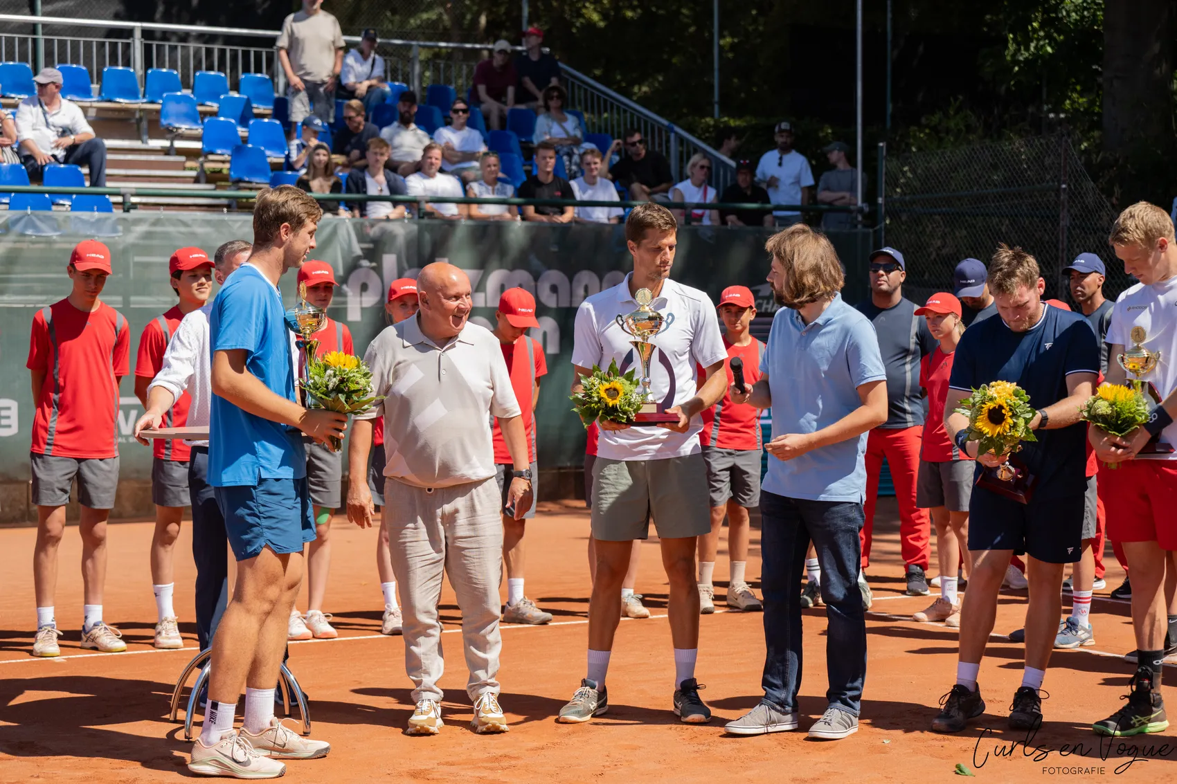 Ein Tennisspieler in einem weißen T-Shirt hält eine Trophäe und einen Sonnenblumenstrauß während einer Preisverleihung auf einem Sandplatz.